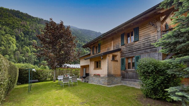 Une maison en bois avec des fondations en pierre, une terrasse avec des chaises et une vue sur une montagne boisée.