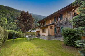 A wooden house with a stone foundation, a patio with chairs, and a view of a forested mountain.