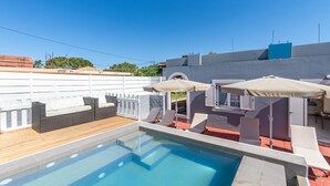 A rooftop swimming pool with lounge chairs and umbrellas, surrounded by a white fence and a building with a blue roof.