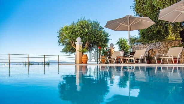 Une piscine d’un bleu éclatant, bordée de chaises longues et d’ombrelles, et entourée de murs de pierre et de verdure.
