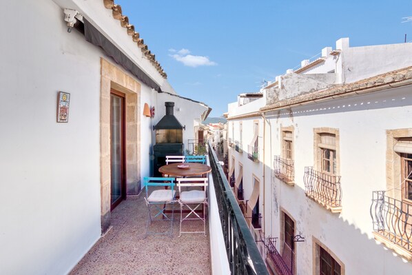 A narrow outdoor balcony with a round table and chairs, a chimney, and white buildings with balconies.