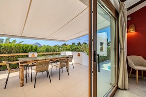 An outdoor dining area with a glass table and wicker chairs, a white wall, and a view of greenery and a mountain. - Holiday Home 'Ca Rulleta Jávea' with Mountain Views, Wi-Fi and Air Conditioning (Jávea)