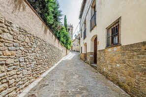 A narrow stone-paved alley with buildings on either side, a stone wall on the left, and a bell tower in the distance.