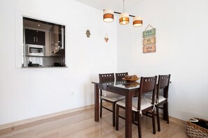 Een eethoek met een tafel, stoelen, een hanglamp en een keuken zichtbaar door een raam.
