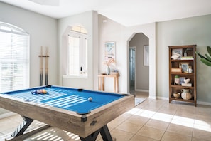 Una mesa de billar en una sala bien iluminada, con una estantería de madera y una puerta que da a otra habitación.