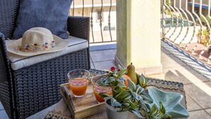 A wicker sofa with a blue cushion and a straw hat on it, a tray with drinks and a potted plant, and a balcony railing.