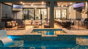 Un espace de vie extérieur moderne doté d’une piscine, de coins salon et d’une cuisine visible à travers de grandes portes vitrées.