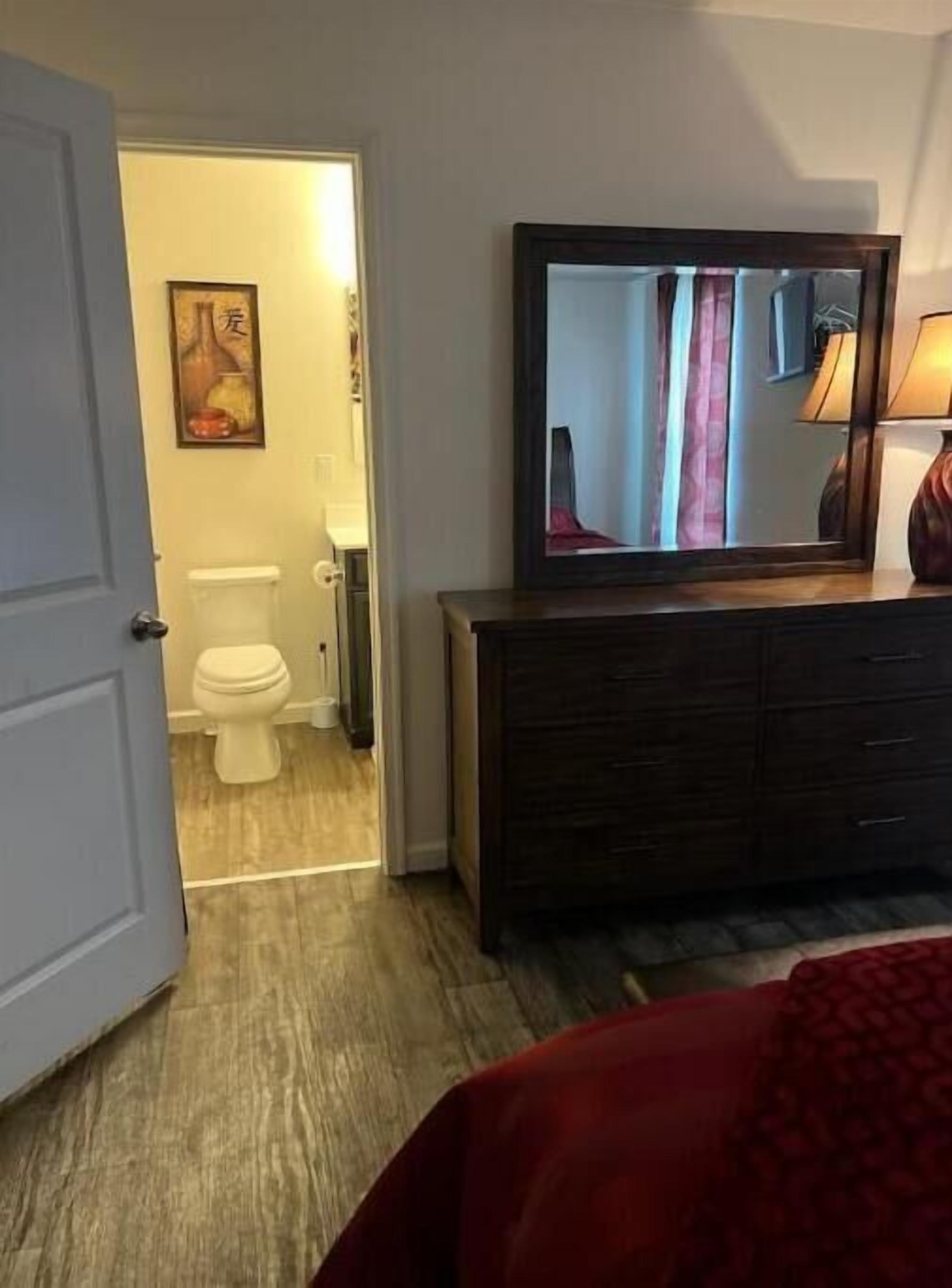 Una habitación de hotel con piso de madera, una puerta blanca, un aparador de madera, un televisor sobre el aparador y un cuadro en la pared.