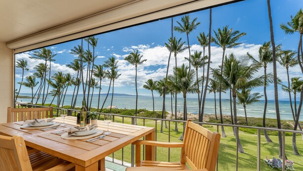 A wooden table and chairs on a balcony with a view of the ocean and palm trees. - Fully Remodeled Ocean Front Comfort HKOK316 (Kihei)