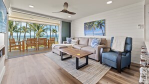 A modern living room with a sofa, armchair, and coffee table, a large window with a view of palm trees and the sea, and a ceiling fan. - Fully Remodeled Ocean Front Comfort HKOK316 (Kihei)