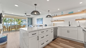 A modern kitchen with white cabinetry, a large island, pendant lighting, and a view of the beach. - Fully Remodeled Ocean Front Comfort HKOK316 (Kihei)