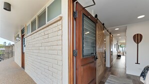 A hallway with a wooden door, a brick wall, and a view into a room with a bed and a wall-mounted decorative item. - Fully Remodeled Ocean Front Comfort HKOK316 (Kihei)