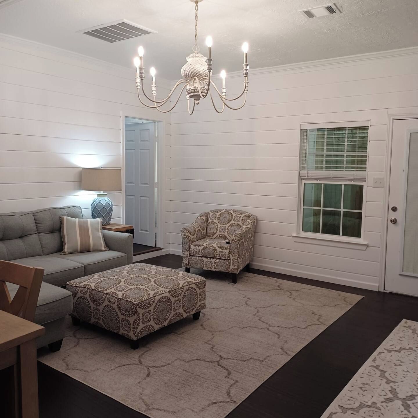 A living room with a sofa, armchair, ottoman, chandelier, and a door with a window.