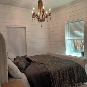 Quarto com cama, lustre, janela com persianas e vista para árvores.