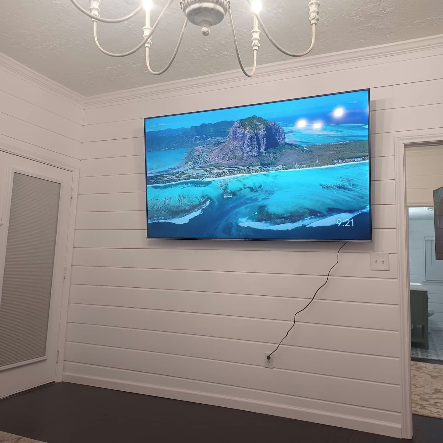 A flat-screen TV mounted on a wall with a landscape image, a chandelier, and a door with a window.