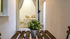 Ein schmaler Flur mit einer hölzernen Bank und einem Blumentopf. Eine Tür führt in einen Raum mit einem Bett und einem Fenster.