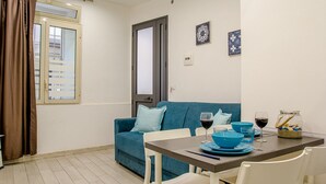 Ein kleiner, moderner Wohnraum mit einem blauen Sofa, einem Esstisch mit blauen Tellern und Gläsern sowie einem Fenster mit Vorhängen.
