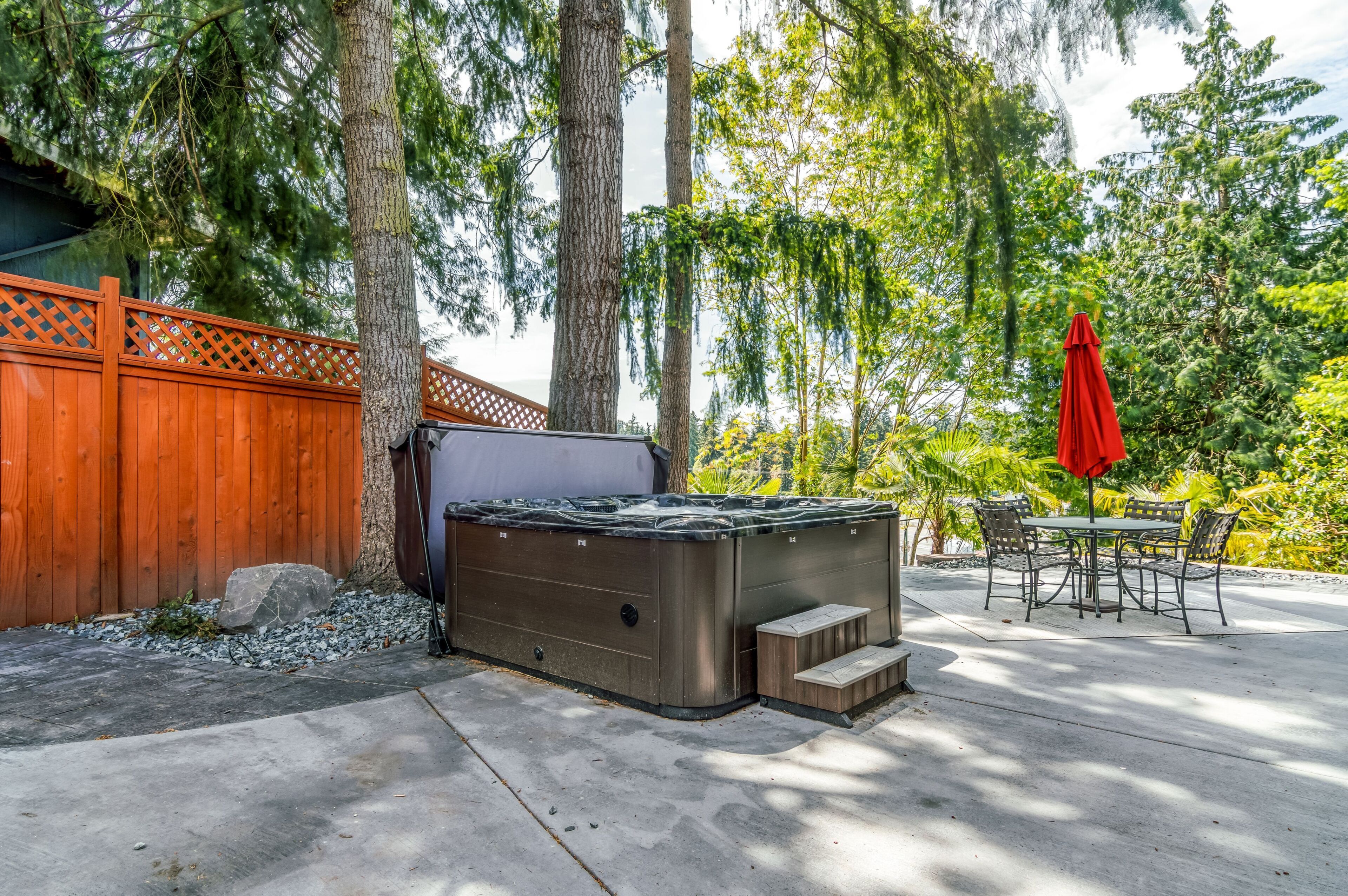 A hot tub in a backyard with a wooden fence, trees, and a patio with chairs and a table.