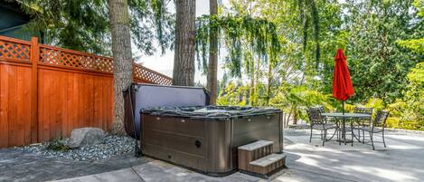A hot tub in a backyard with a wooden fence, trees, and a patio with chairs and a table.