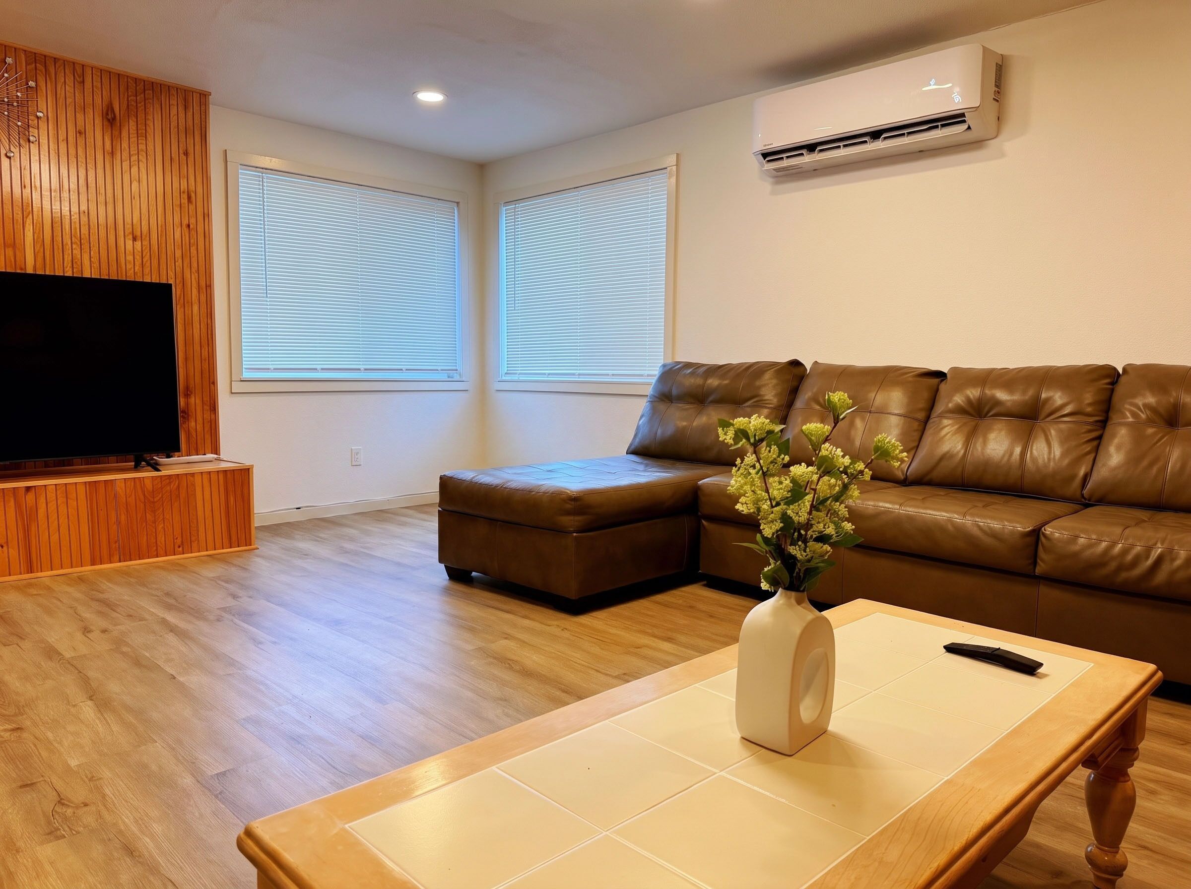 Een woonkamer met een televisie, een bruine leren bank, een witte vaas met bloemen en een houten salontafel.