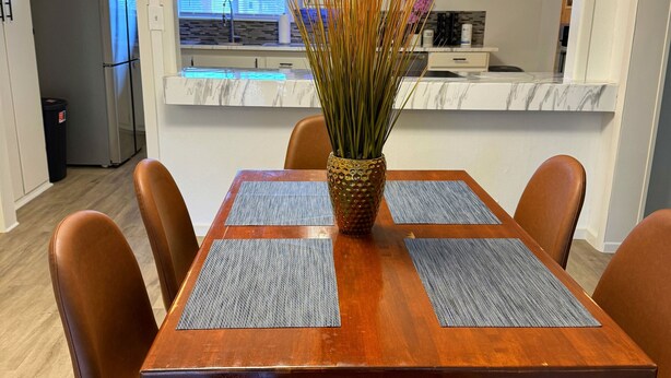 Un coin repas avec une table en bois, quatre chaises marron et un vase décoratif contenant de hautes herbes.
