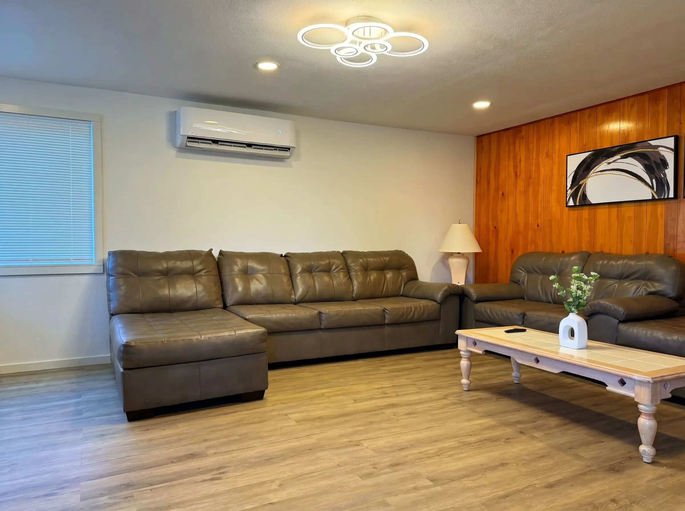 A living room with a sectional sofa, a coffee table, an air conditioning unit, and a wooden wall panel.