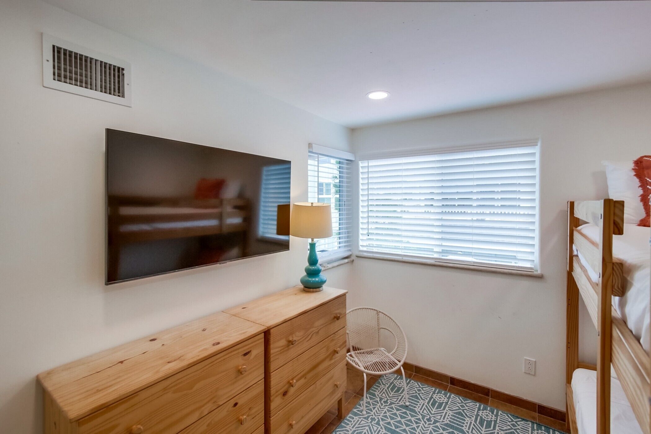A bedroom with a wooden dresser, a chair, a bunk bed, a TV, and a window with blinds.