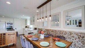 Une cuisine moderne avec des meubles blancs, une table à manger en bois mise avec des assiettes et des verres, et des luminaires suspendus au-dessus.
