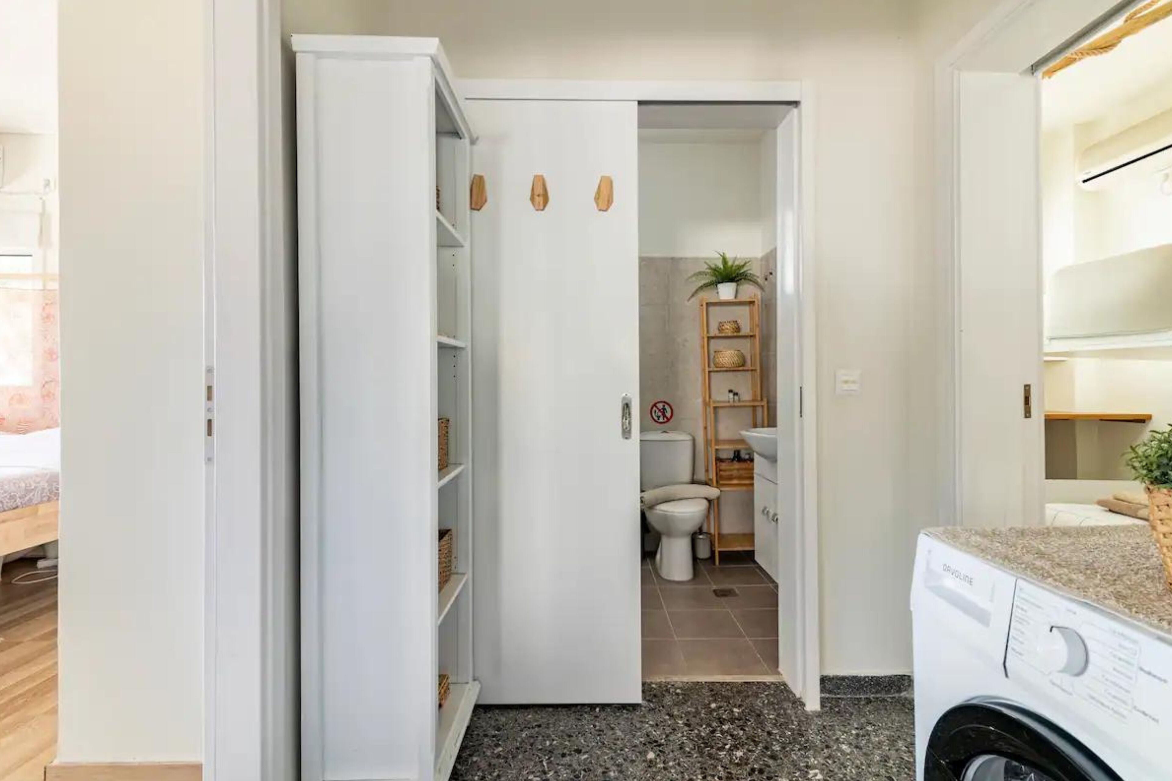 A laundry room with a washing machine, a shelf with baskets, and a toilet visible through an open door.