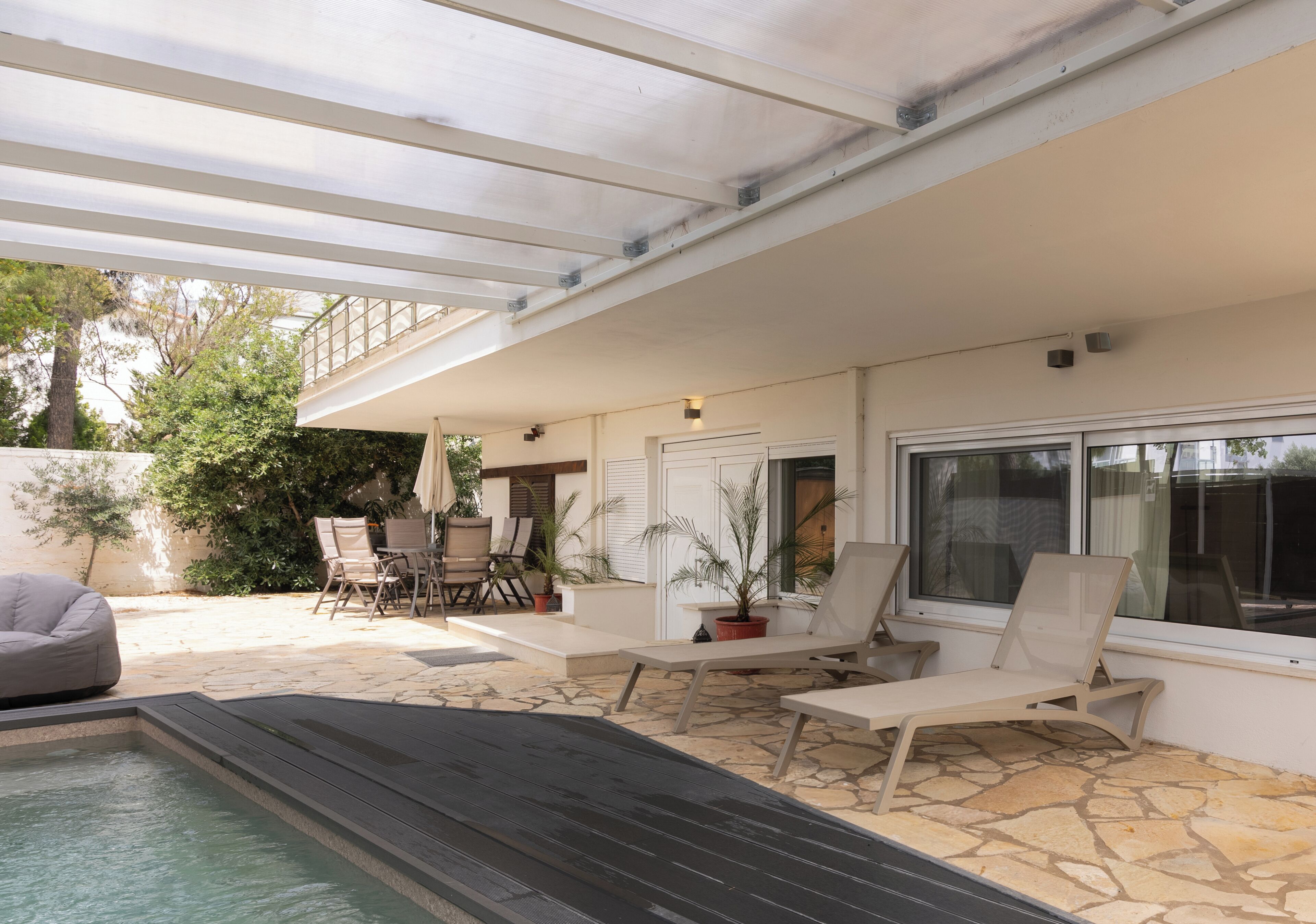 Un espacio exterior techado con piscina, sillas de descanso y una mesa con sillas.