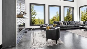 A modern living room with a fireplace, large windows, and a view of trees. - Knife River Cabin with Amazing View and Sauna (Two Harbors)