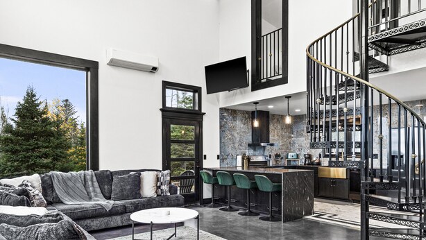 A modern living room with a large sofa, a coffee table, and a unique gold dripping table. A staircase with a black railing leads to an upper level. - Knife River Cabin with Amazing View and Sauna (Two Harbors)