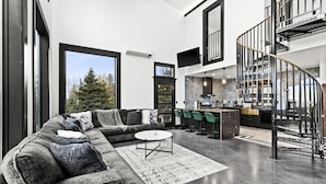 A modern living room with a sectional sofa, a round coffee table, and a view of the kitchen and staircase. - Knife River Cabin with Amazing View and Sauna (Two Harbors)