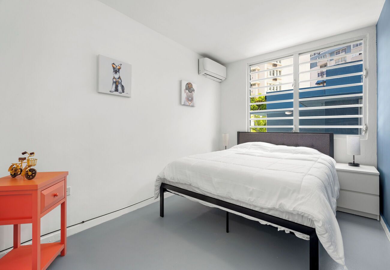 A bedroom with a bed, a nightstand, a window with blinds, and a small table with a toy bicycle.