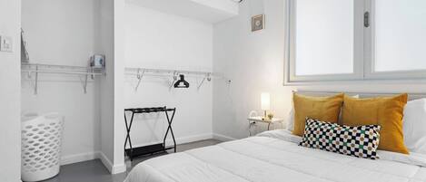 A modern bedroom with a large bed, a window, a wall-mounted air conditioner, a laundry basket, and a small table with a lamp.