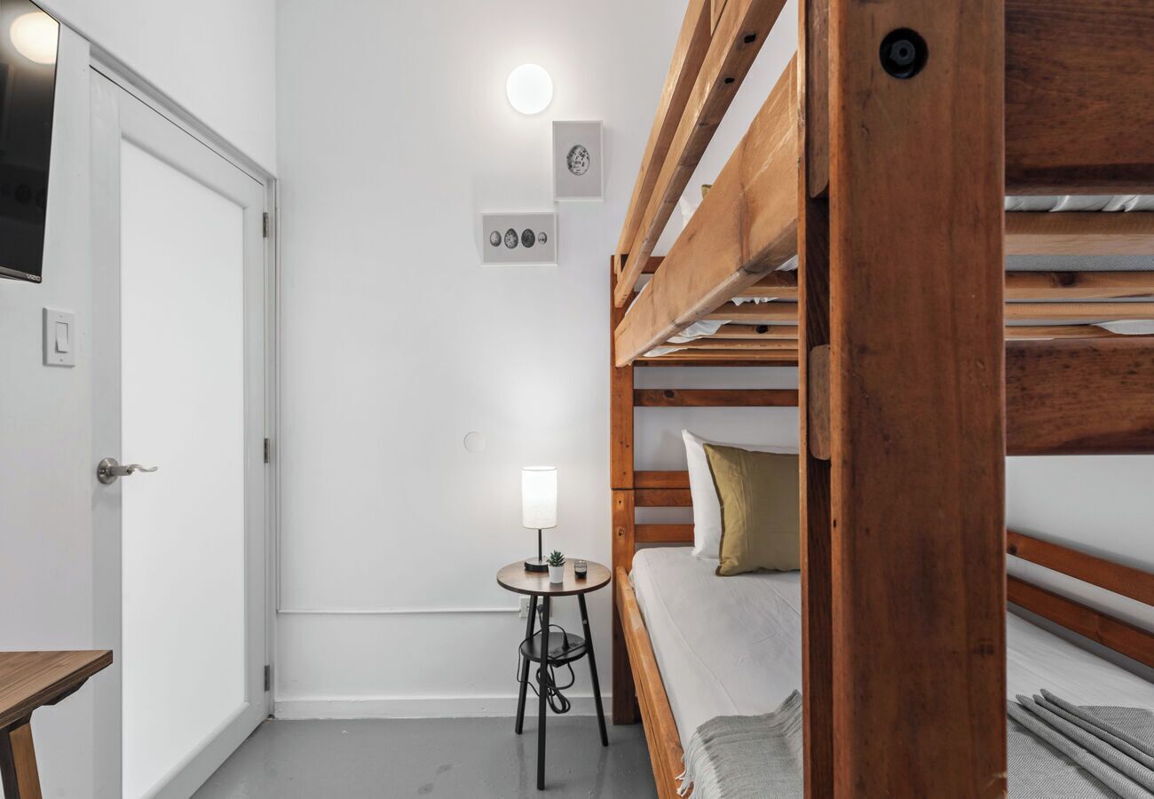 Dormitorio con literas, una mesita pequeña y una lámpara, paredes blancas y una puerta.
