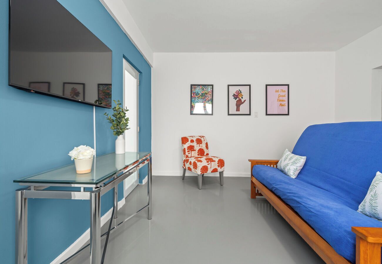 Un salon moderne avec un canapé bleu, une table basse en verre, un fauteuil à motifs floraux rouges et un téléviseur fixé au mur.