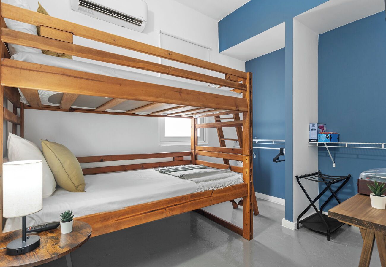 Un lit superposé avec une structure en bois, une petite table avec une lampe et une plante, et un bureau avec une chaise et une plante en pot.