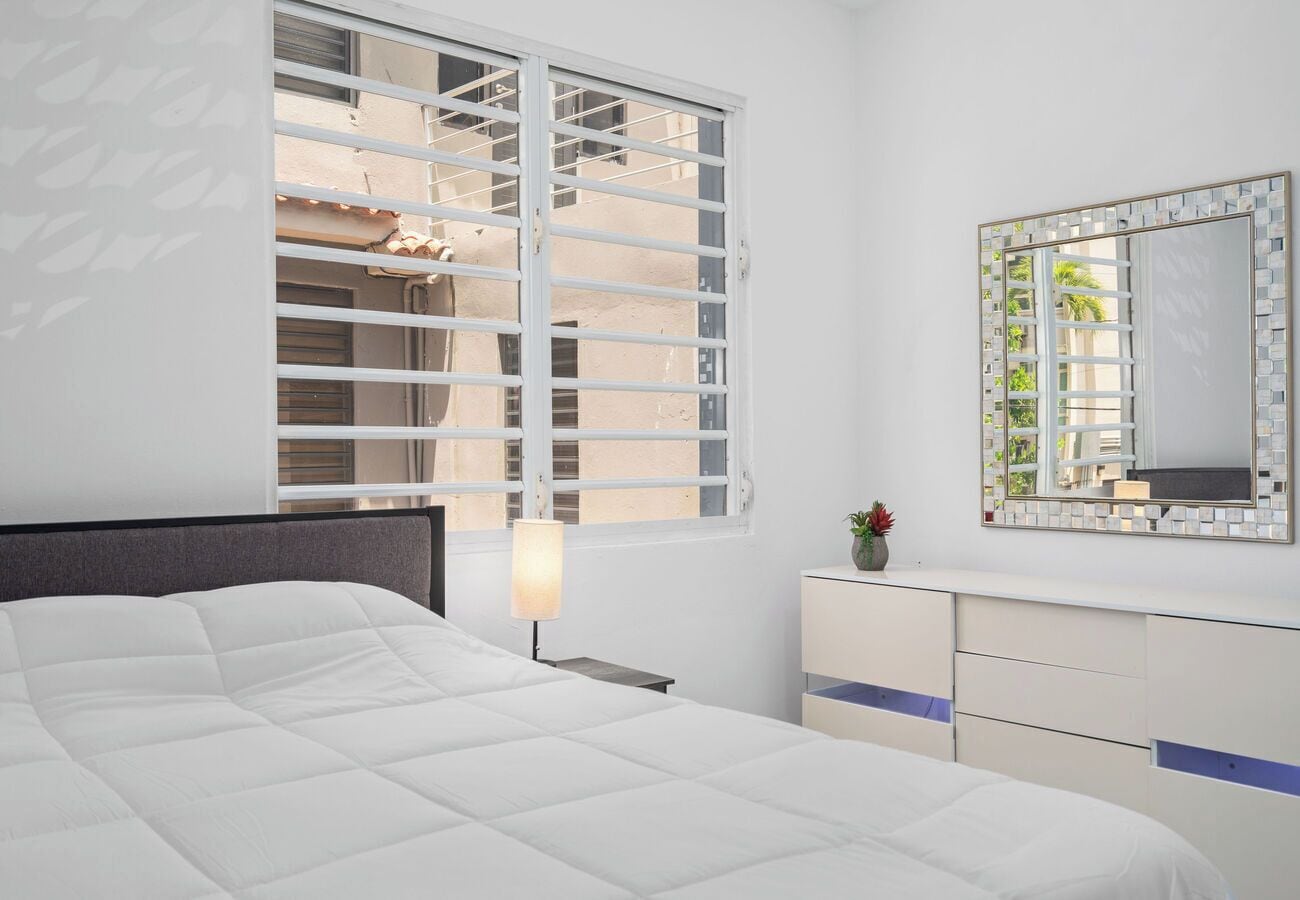 A bedroom with a bed, a nightstand, a dresser, a mirror, and a window with blinds.