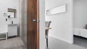 Una habitación moderna y minimalista con puerta de madera, lavamanos blanco, espejo y cama con edredón blanco.