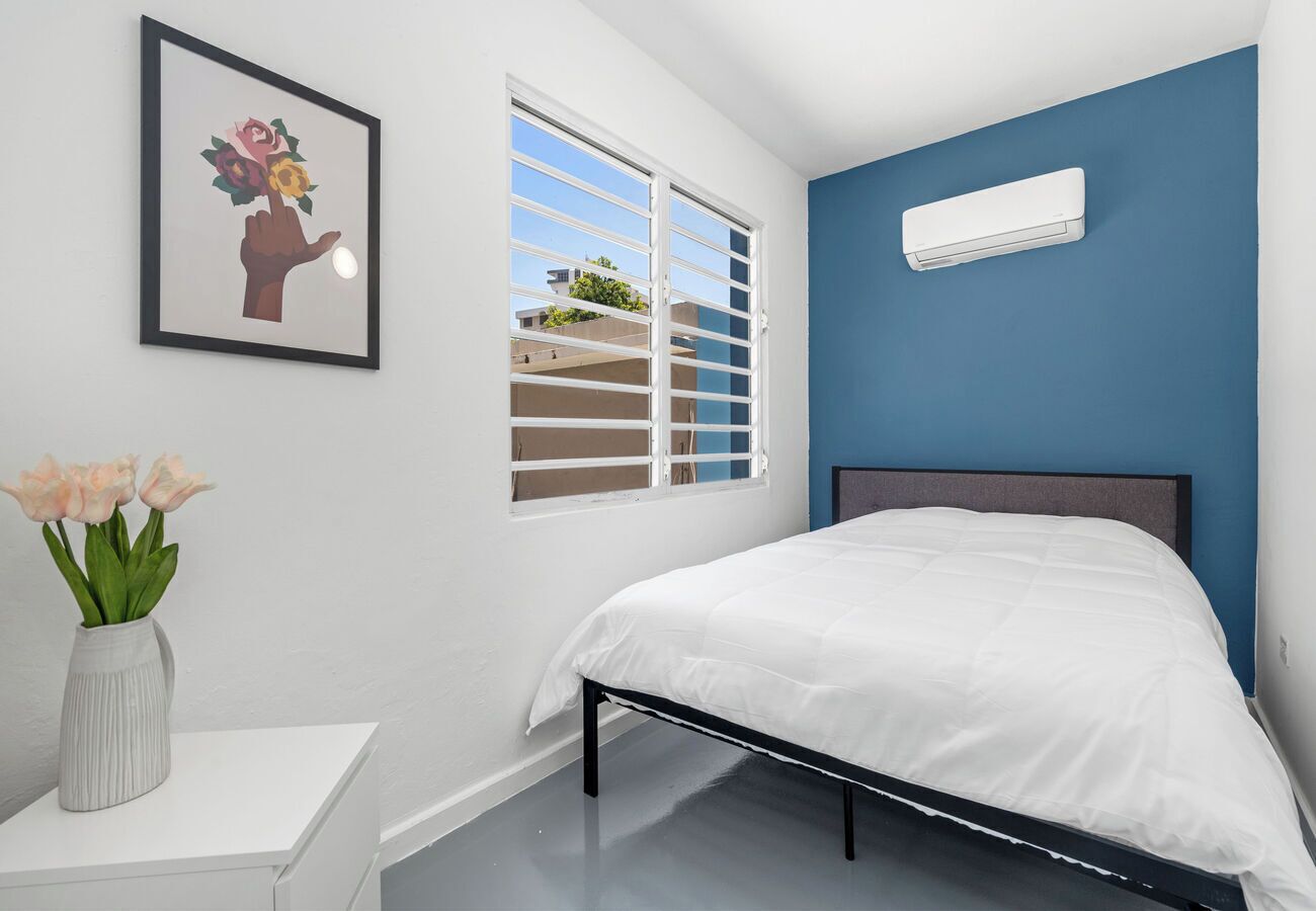 Ein Schlafzimmer mit Bett, einem Fenster mit Jalousien, einer Wand-Klimaanlage und einer Blumenvase auf einem Nachttisch.