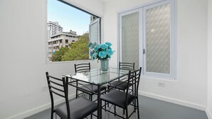 Un comedor con mesa de vidrio y sillas negras, un florero con flores azules, y vista a edificios y árboles a través de grandes ventanales.