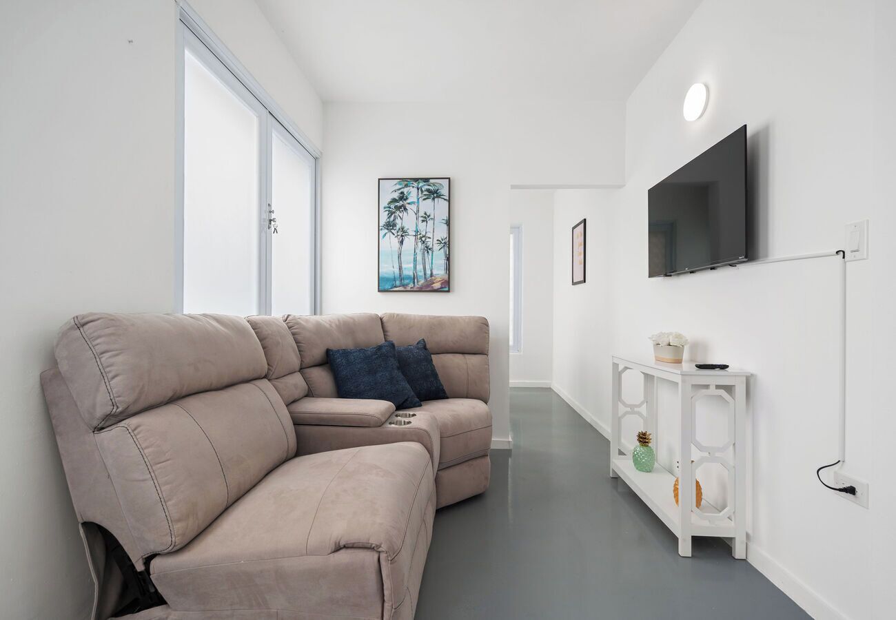 A modern living room with a beige sectional sofa, a white console table, a flat-screen TV mounted on the wall, and a painting of palm trees.