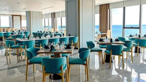 A spacious restaurant with teal chairs and tables, large windows offering a view of the sea, and a polished marble floor. - Apec Mandala Beach Mui Ne Resort (Mui Ne)