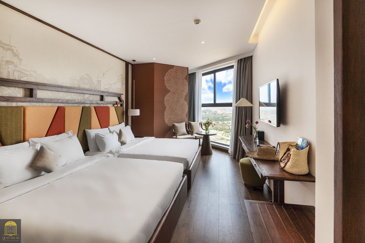 A hotel room with two beds, a desk, a chair, a TV, and a window with a view.