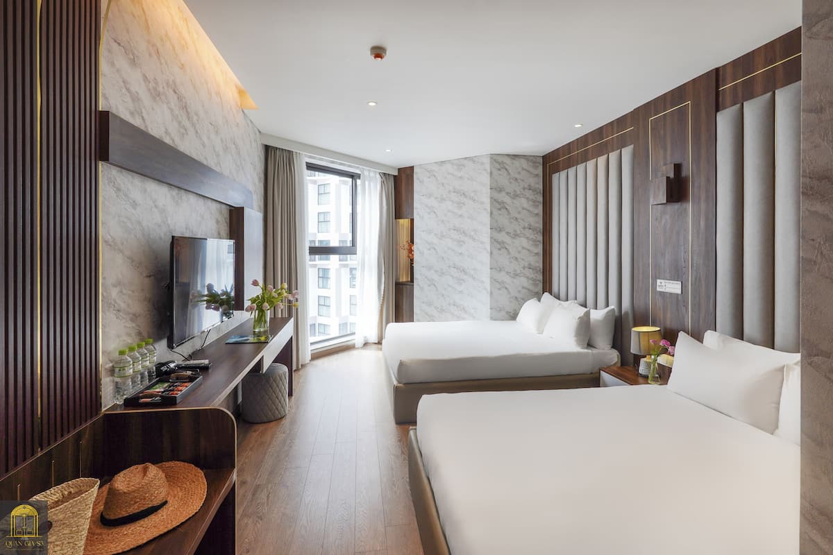 A hotel room with a large bed, a smaller bed, a TV, a desk with a computer, and a window with a view of buildings.