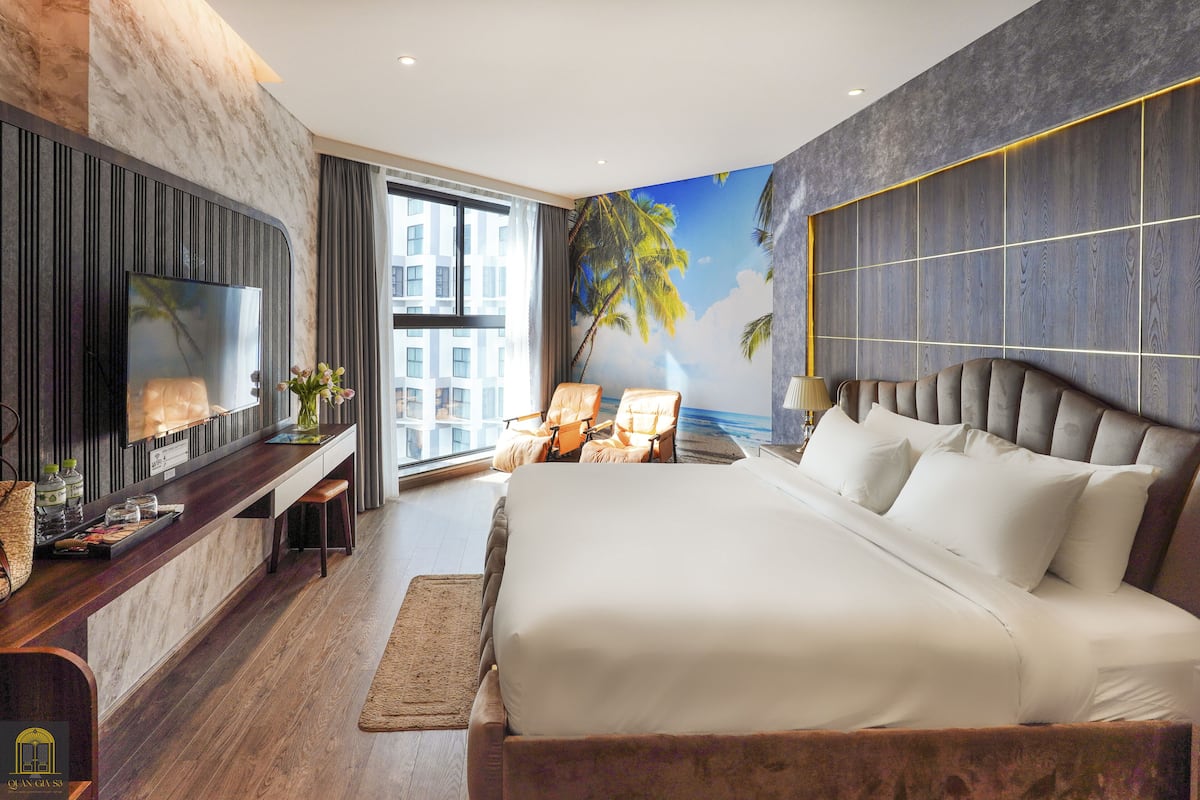A modern hotel room with a large bed, a TV mounted on the wall, a seating area with chairs, and a mural of palm trees.