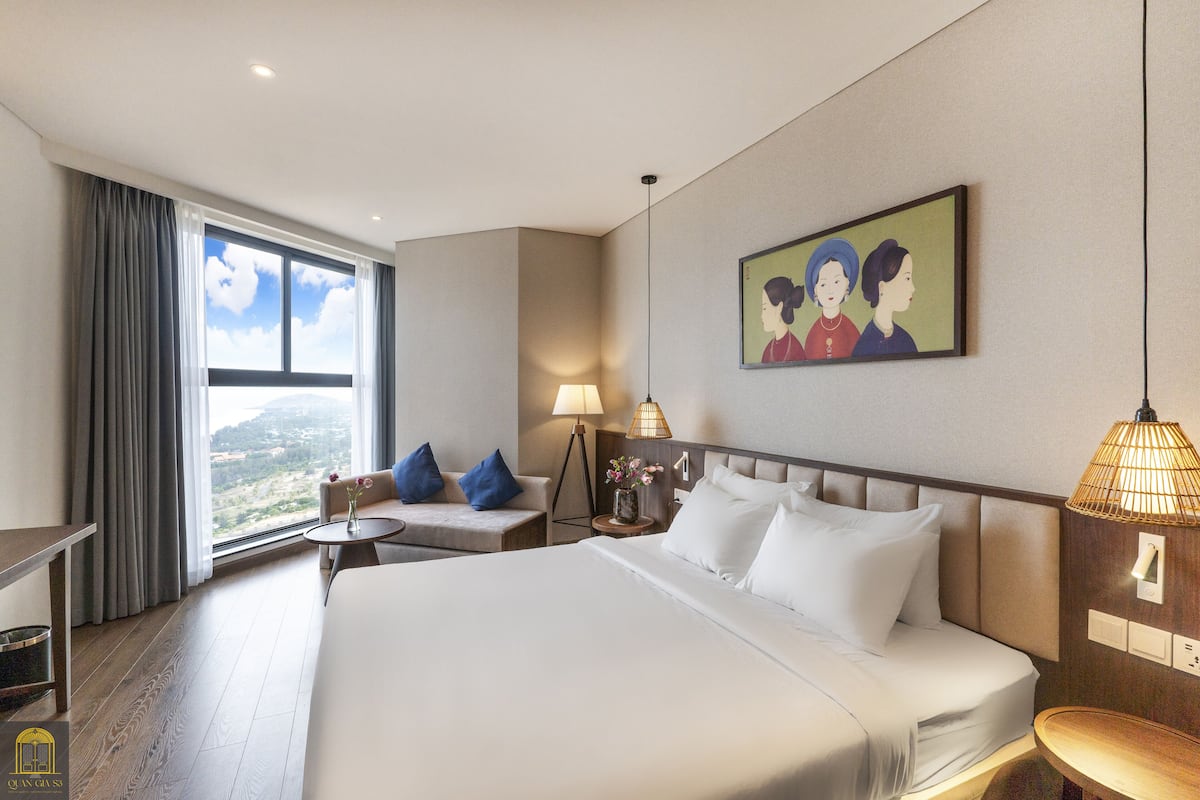 A hotel room with a large bed, a desk, a chair, a small table, a lamp, and a painting on the wall.