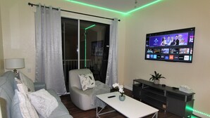 Una sala de estar con un sofá gris, una mesa de centro blanca, un televisor montado en la pared y una puerta corrediza de vidrio con cortinas.