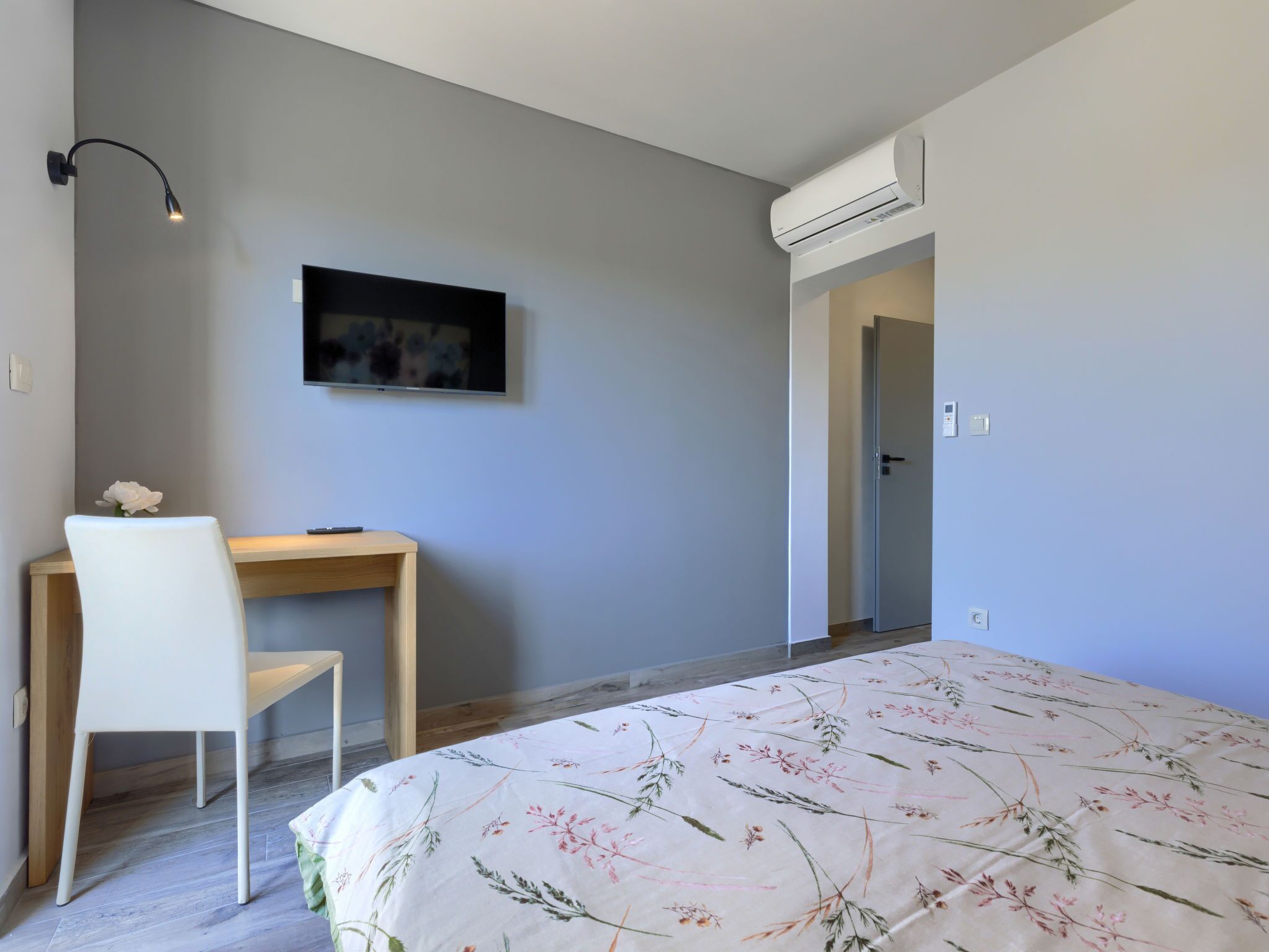 A bedroom with a bed, a desk, a chair, a wall-mounted air conditioner, and a door leading to another room.